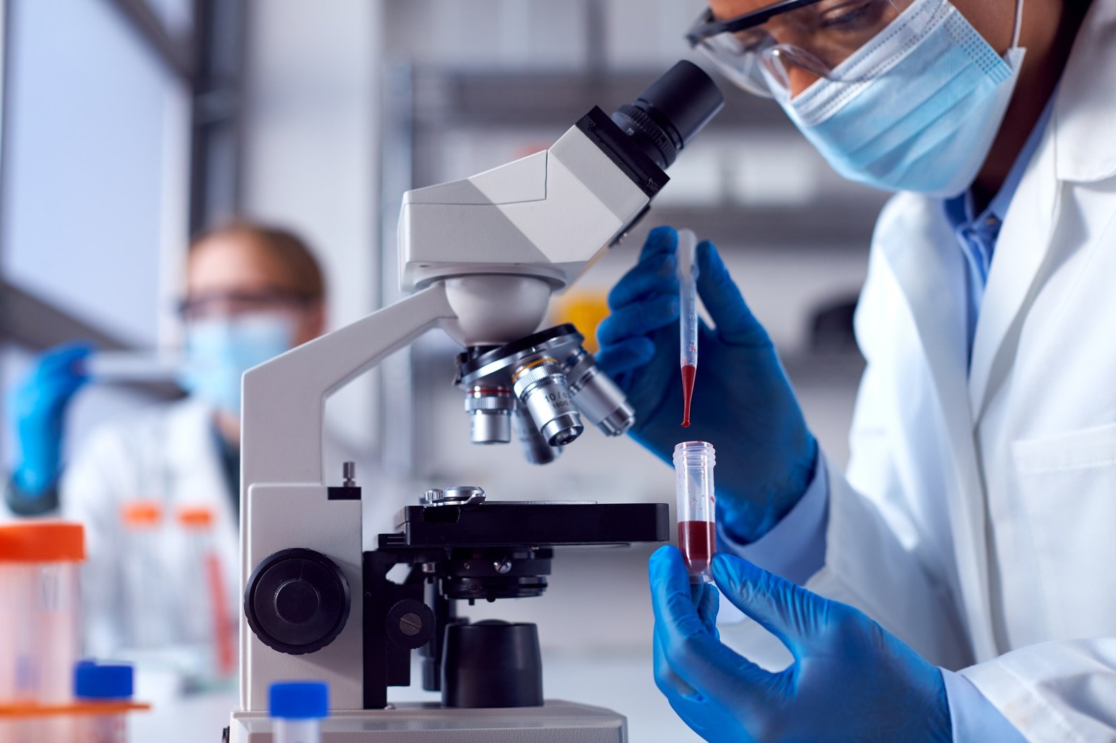 Two Female Lab Workers Wearing PPE Testing Blood Samples In Laboratory With Microscope